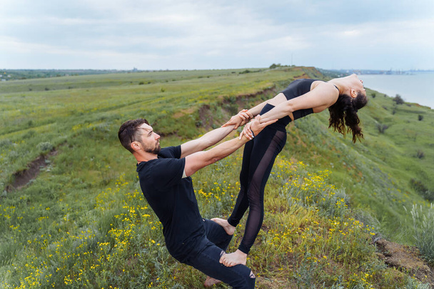 Acro sport. Professional athletes in black sportswear balancing perfectly on the edge of hill against of the sea. Sportsman holding himself and the girl on his feet, leaning back.  - Photo, Image