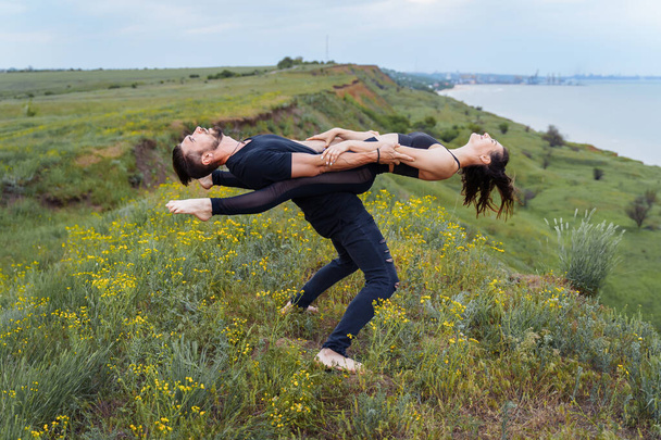 Acro sport. Professional athletes in black sportswear balancing perfectly on the edge of hill against of the sea. Sportsman holding himself and the girl on his feet, leaning back. They create line - Photo, Image