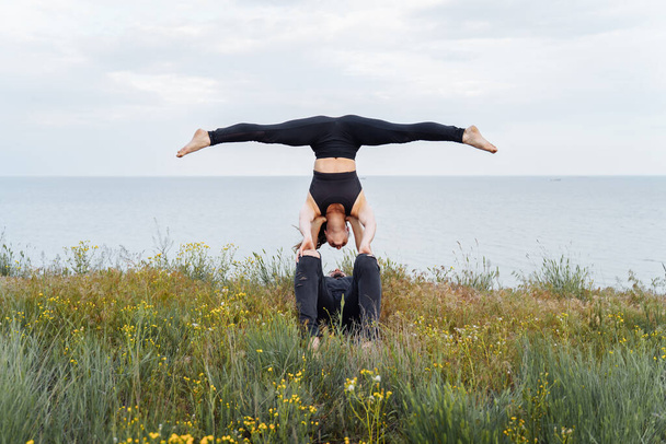 Atraktivní pár cvičit jógu venku na břehu moře. Děvče dělá rozchody, je vzhůru nohama během akroyogy, leží na nohou muže. Sportovní cvičení na kopci. Vzdálený výstřel - Fotografie, Obrázek