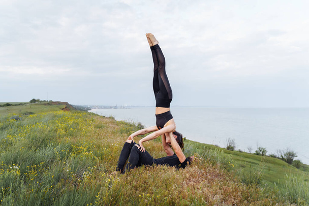Two young sportsmen doing fitness acro yoga exercise together outdoors. Acrobatic elements for strength and balance at sea on the hill.  Man lifting and balancing woman. Healthy lifestyle - Photo, Image