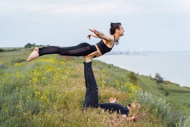 Young couple guy and girl doing acro yoga on the background of the sea. Man lying on the grass and balancing woman on his legs. Dressed in black sportswear. Woman is flying in the air - Photo, Image