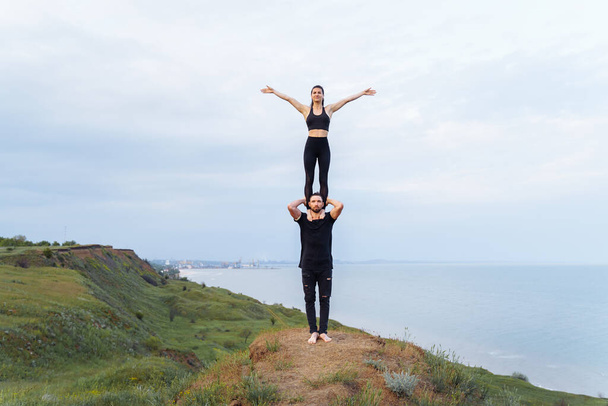 Strong, muscular athlete holding on his shoulders a fit, beautiful sportswoman girl. Acrobats warming up before training, standing on cliff hill on the background of the sea. Tricks akroyoga in pairs - Photo, Image
