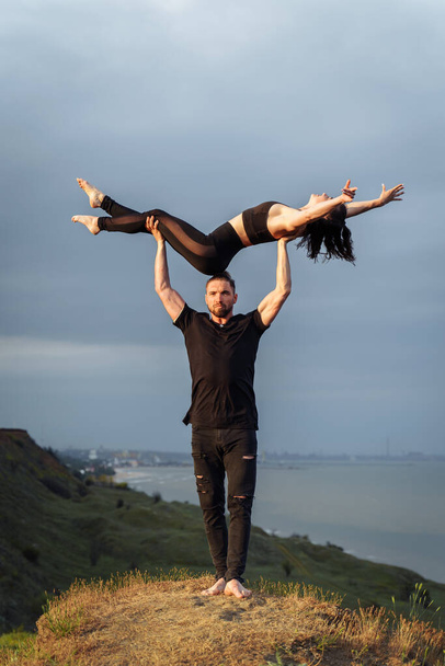 Full-length portrait. Attractive muscular, pumped-up young man is doing in acroyoga with his girlfriend. Instead of a bar, he lifts her up on outstretched arms and smiles. Standing on the edge of hill - Photo, Image