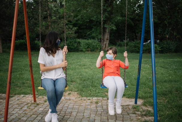 Woman and little girl on swing while wearing antivirus masks - Foto, immagini