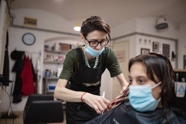 Kapper en meisje kind klant in een salon met medische maskers tijdens virus pandemie. Werken met veiligheidsmasker. - Foto, afbeelding