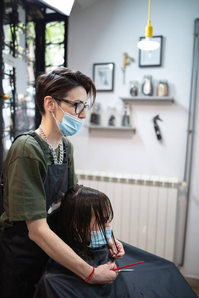 Peluquería y niña cliente en un salón con máscaras médicas durante la pandemia de virus. Trabajar con máscara de seguridad
. - Foto, Imagen