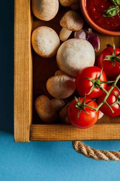 vista superior de la salsa de tomate en tazón cerca de setas, tomates rojos cereza y dientes de ajo en caja de madera en azul
 - Foto, Imagen