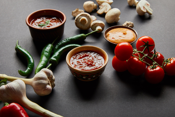 selective focus of tasty sauces in bowls near ripe cherry tomatoes, green chili peppers, mushrooms and garlic on black - Zdjęcie, obraz