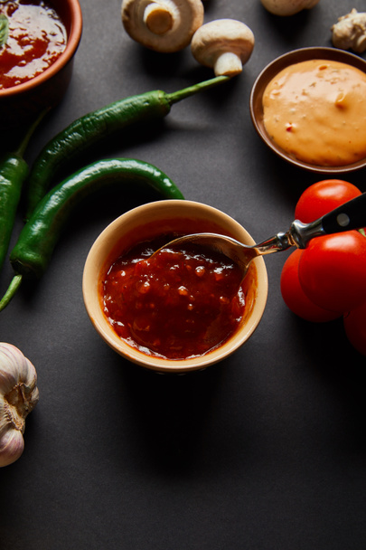 top view of tasty sauces in bowls near ripe cherry tomatoes, green chili peppers and mushrooms on black - Фото, зображення