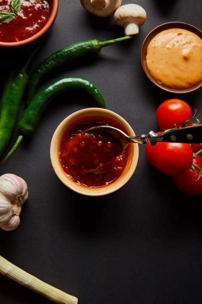 top view of tasty sauces in bowls near ripe cherry tomatoes, garlic, green chili peppers and mushrooms on black - Φωτογραφία, εικόνα