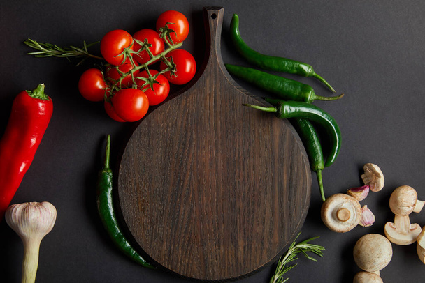 top view of wooden cutting board near ripe cherry tomatoes, garlic, rosemary, green chili peppers and mushrooms on black - Фото, изображение