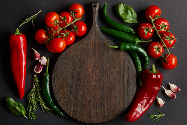 top view of wooden chopping board near ripe cherry tomatoes, garlic cloves, rosemary, basil leaves and green chili peppers on black - Фото, изображение