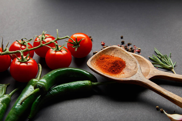 red cherry tomatoes, rosemary, peppercorns, wooden spoons with paprika powder and green chili peppers on black - Photo, Image