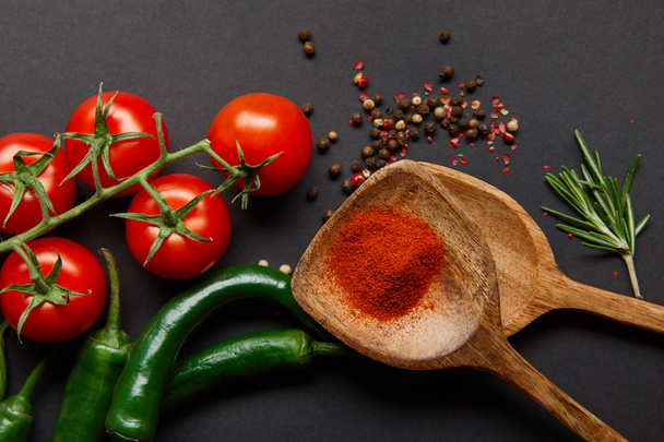 vista superior de tomates rojos cereza, romero, granos de pimienta, cucharas de madera con pimentón en polvo y chiles verdes en negro
 - Foto, Imagen