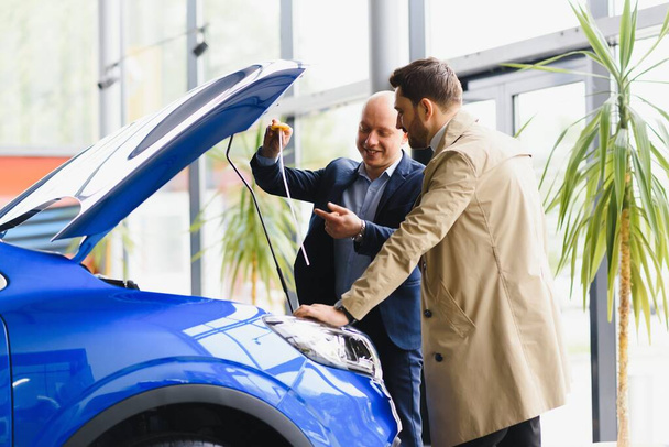 Car dealer and his ale client looking under the hood of a new car on sale at the dealership. Young man buying auto at car salon - 写真・画像