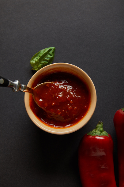 Верхний вид ложки в миске с томатным соусом возле листьев базилика и перца чили на черном
  - Фото, изображение