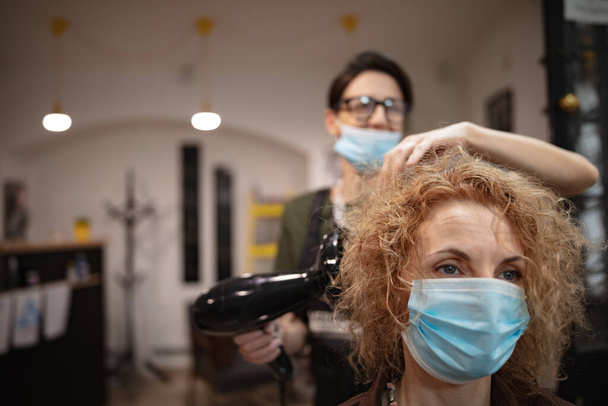 Peluquería y cliente en un salón con máscaras médicas durante la pandemia de virus. Trabajar con máscara de seguridad. - Foto, imagen