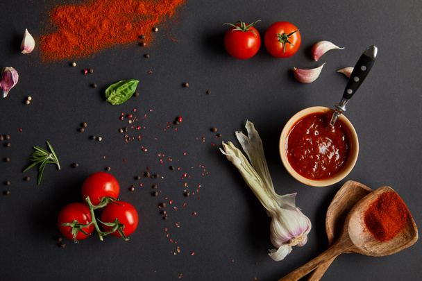 vista superior de tomates rojos cereza, dientes de ajo y hierbas frescas cerca de cucharas de madera con pimentón en polvo sobre negro
  - Foto, imagen