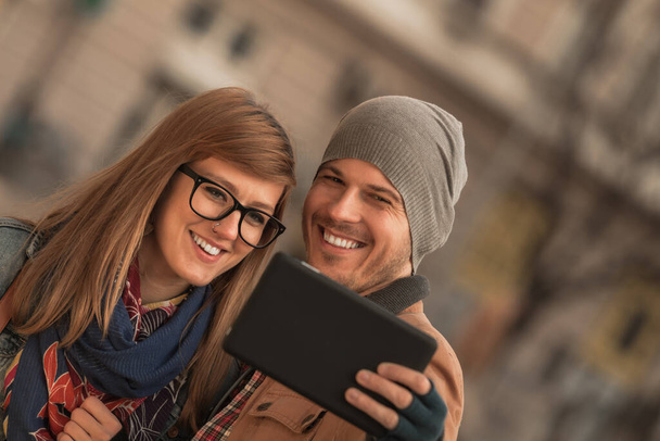 Νεαρό ζευγάρι τουριστών που χρησιμοποιούν έξυπνο tablet στο αστικό περιβάλλον της πόλης για τη λήψη φωτογραφιών. - Φωτογραφία, εικόνα