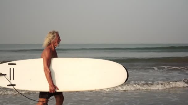 Starší surfař. Šedovlasý děda na pláži s prknem - Záběry, video