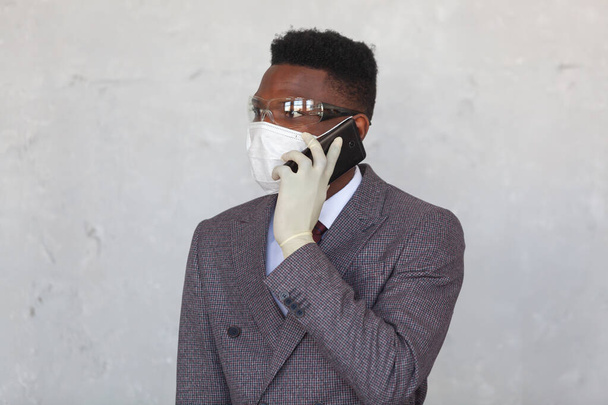 COVID-19. Handsome African black man in a suit is using medical mask, protective glasses and holding his mobile phone with a hand in gloves. Measures to prevent coronavirus infection and the - Fotografie, Obrázek