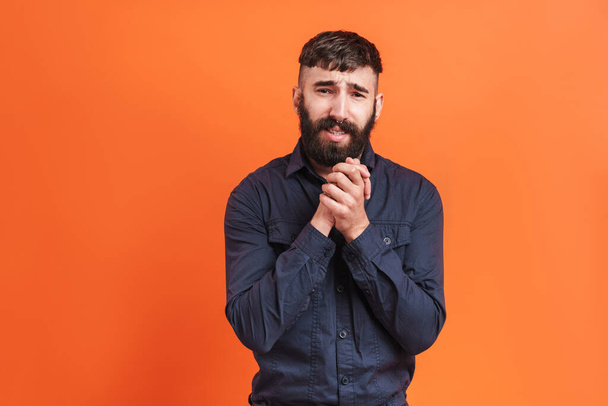 Image closeup of uptight man with nose jewelry wearing black shirt praying and keeping hands together isolated over orange background - Foto, Imagen