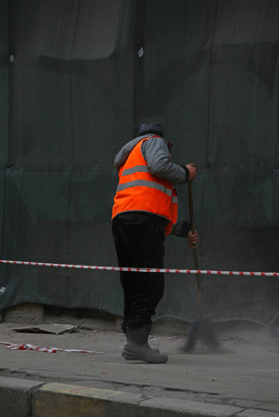 údržbář v oranžové vestě zametá chodník s koštětem vedle budovy pokryté ochrannou sítí - Fotografie, Obrázek
