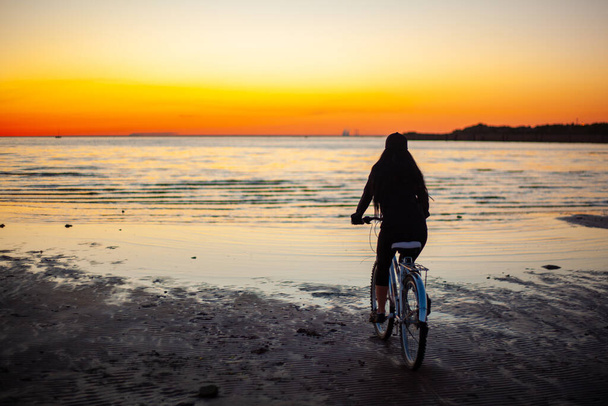 後ろから海で自転車に乗っている少女のシルエット。日の出. - 写真・画像