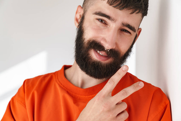 Image of handsome bearded man with nose jewelry wearing orange shirt showing peace sign while standing over white wall indoors - Photo, image