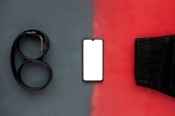 Ropa y accesorios modernos para mujer. Vista superior del cinturón de cuero negro de moda, jeans negros y teléfono sobre fondo gris y rojo
. - Foto, imagen