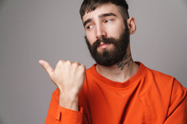Image closeup of young bearded man with nose jewelry wearing orange shirt pointing finger at copyspace isolated over gray background - 写真・画像