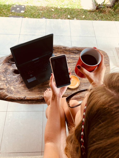Modern freelance young woman using cellphone / laptop and drinking coffee on a terrace sofa. - Photo, Image