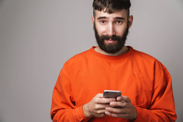 Image closeup of young bearded man with nose jewelry wearing orange shirt holding smartphone isolated over gray background - Foto, Imagem