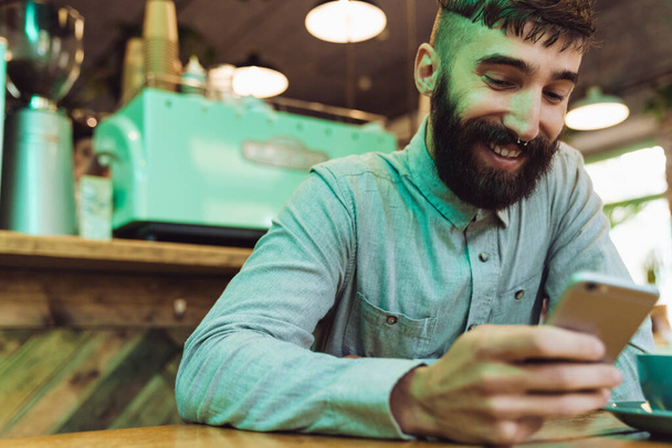 Attractive smiling young bearded man wearing shirt using mobile phone while sitting at the cafe indoors and drinking coffee - Foto, afbeelding