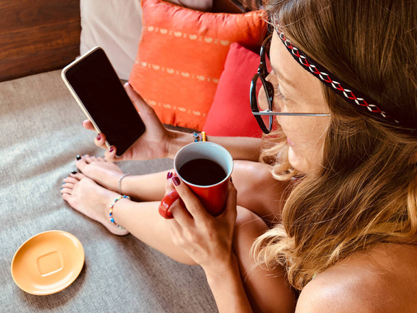 Сучасна молода жінка використовує мобільний телефон і п'є каву на терасному дивані
. - Фото, зображення
