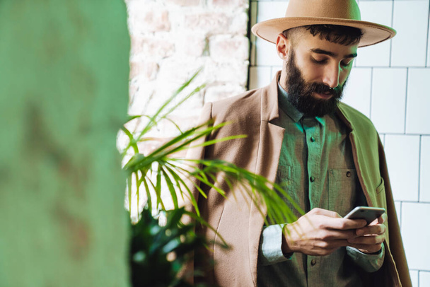 Attractive young bearded man wearing coat using mobile phone while sitting at the cafe indoors at the window - Foto, imagen