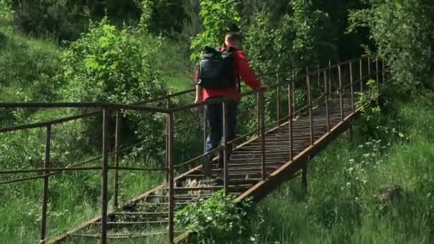赤いジャケットと黒いバックパックを着た若い男が放棄された場所に移動します。彼は夏の日に森の古い錆びた階段を登り. - 映像、動画