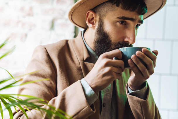 Image of attractive stylish young man wearing hat drinking tea from cup in cafe - Foto, Bild
