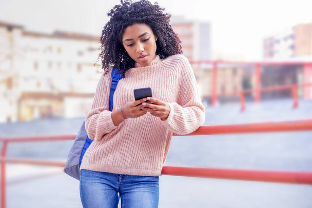 Siyahi öğrenci kız şehir arka planında cep telefonuyla mesajlaşıyor. - Fotoğraf, Görsel