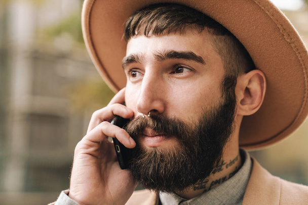 Image of attractive stylish young man wearing hat talking on cellphone while walking outdoors - Fotografie, Obrázek