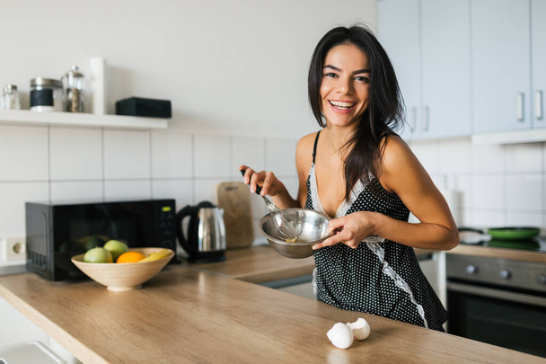 portrait of young brunette pretty woman cooking scrambled eggs in kitchen in morning, smiling, happy mood, positive housewife, healthy lifestyle, laughing, having fun - Foto, Bild