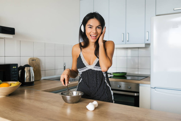 portrait of young brunette attractive woman cooking scrambled eggs in kitchen in morning, smiling, happy mood, positive housewife, healthy lifestyle, listening to music on headphones - Фото, изображение