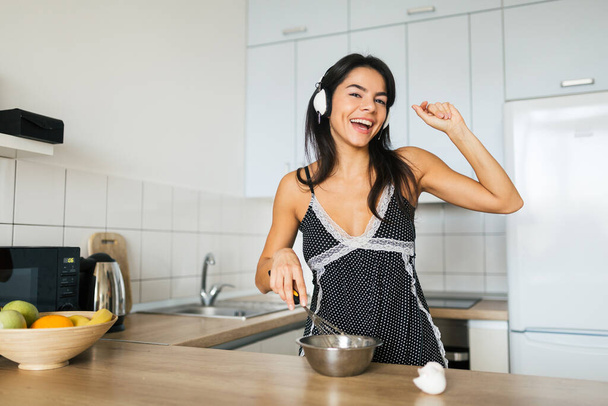 young attractive woman cooking scrambled eggs in kitchen in morning, smiling, happy positive housewife, healthy lifestyle, listening to music on headphones, laughing, having fun, dancing, singing - Foto, imagen