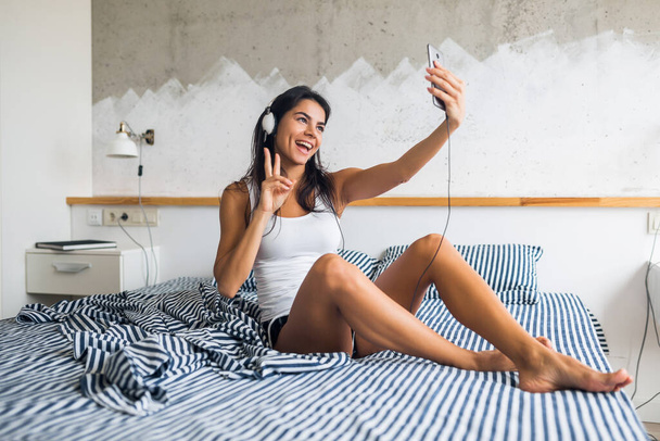 mladá atraktivní žena sedící na posteli v pyžamu, usmívající se v ložnici, šťastná emoce, ráno vstávat, smát se, sexy, hubená, poslouchat hudbu na sluchátkách, používat smartphone, fotit selfie - Fotografie, Obrázek