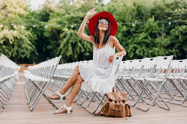 ελκυστική γυναίκα ντυμένη με λευκό φόρεμα, κόκκινο καπέλο, γυαλιά ηλίου που κάθεται στο θερινό υπαίθριο θέατρο στην καρέκλα και μόνο, άνοιξη δρόμο μόδα τάση, εξαρτήματα, ταξιδεύουν με σακίδιο - Φωτογραφία, εικόνα