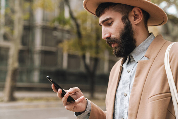 Image of attractive stylish young man wearing hat holding cellphone while walking outdoors - Fotó, kép