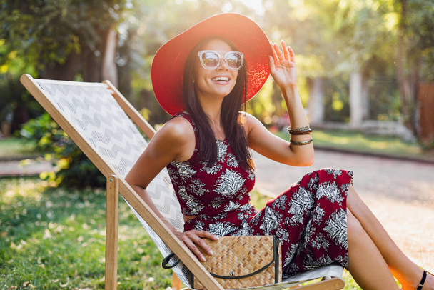 熱帯の衣装を着た美しい女性がデッキチェアに座っています。ストリートスタイルの夏のファッショントレンドの女性。生地のハンドバッグ赤い帽子サングラス。休暇中の幸せな気分で微笑むスタイリッシュな女の子. - 写真・画像