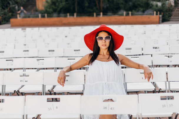 attractive happy smiling woman dressed in white dress, red hat, sunglasses sitting in summer open air theatre alone, many chairs, spring street style fashion trend - Foto, afbeelding