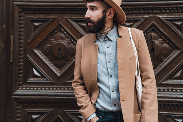 Attractive young man wearing autumn coat and hat standing outdoors at the city street in front of the wooden door - Foto, imagen
