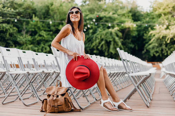 attractive woman dressed in white dress, red hat, sunglasses sitting in summer open air theatre on chair alone, spring street style fashion trend, accessories, traveling with backpack - 写真・画像